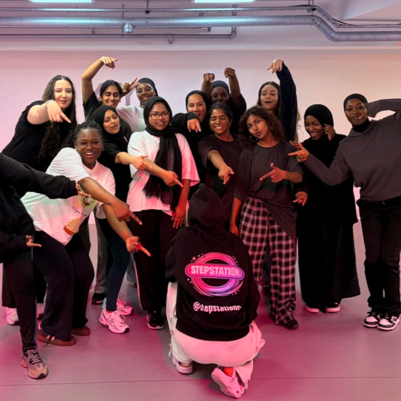 A group of young women pose for a photo in a dance studio with pink and white lighting.
