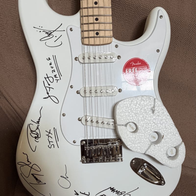 A white electric guitar covered in signatures rests on a brown textured surface.