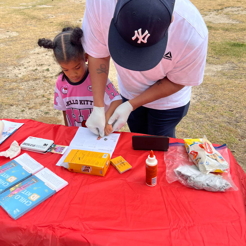 A person in a white shirt and baseball cap, wearing gloves, helps a young girl in a pink Hello Kitty shirt take a fingerprint on a table covered with a red tablecloth, with a grassy field in the background.