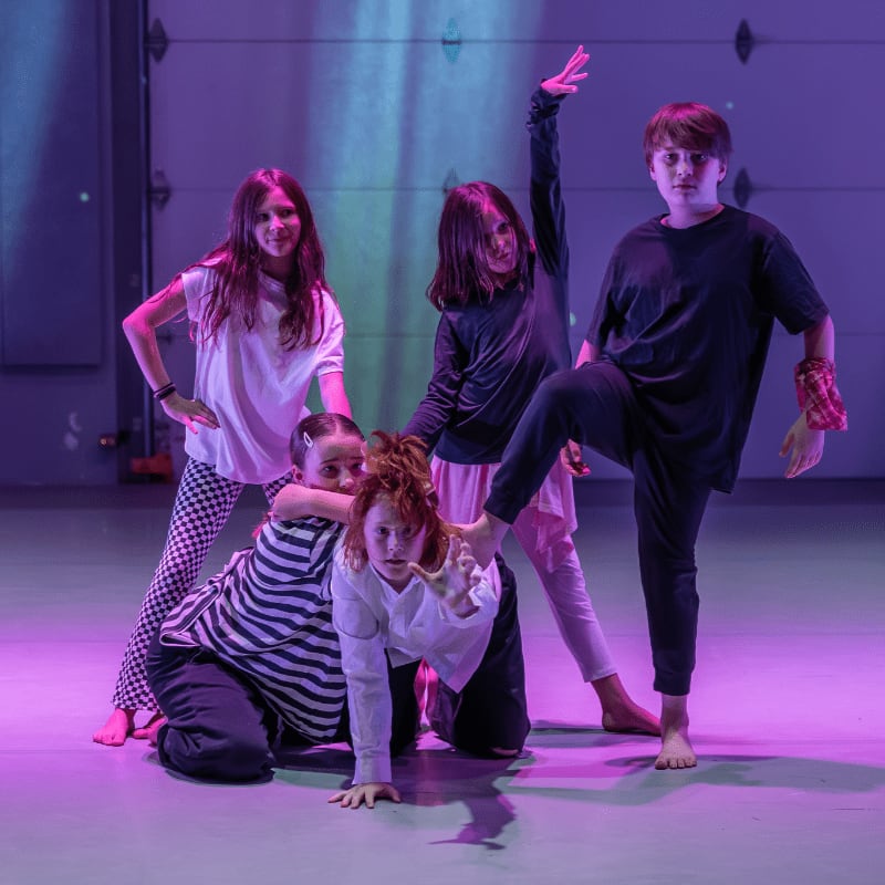 Five children in dynamic poses are illuminated by purple stage lights against a blurred garage door background.