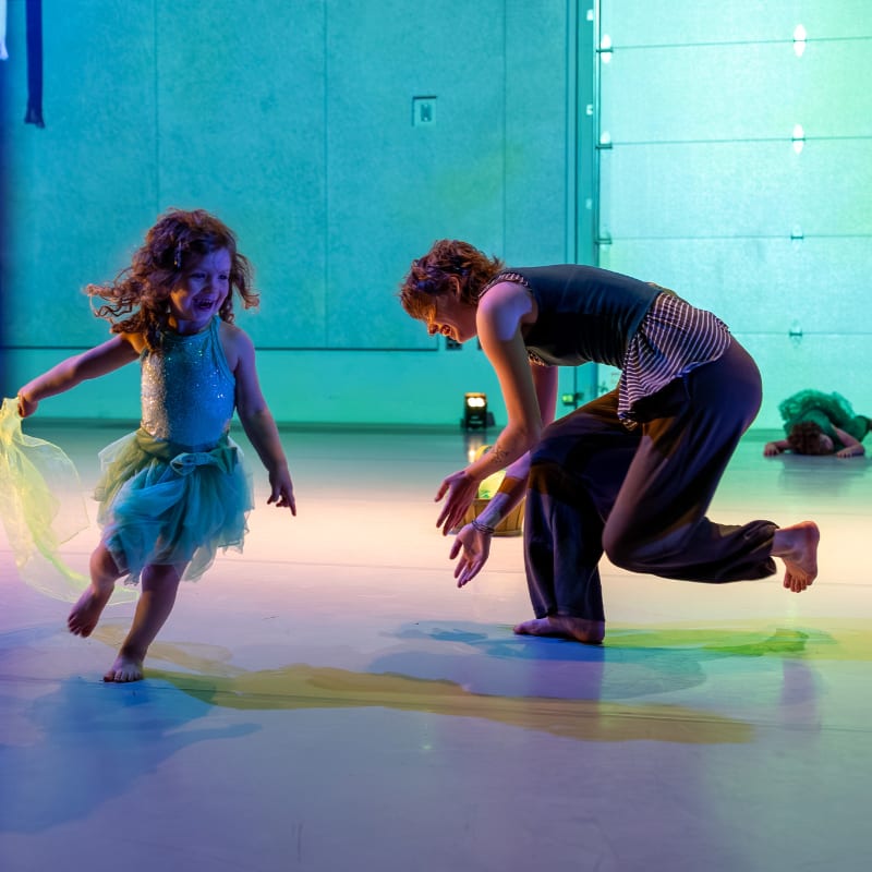 A young girl in a sparkly dress dances with an adult on a colorful stage, while another person lies on the floor in the background.