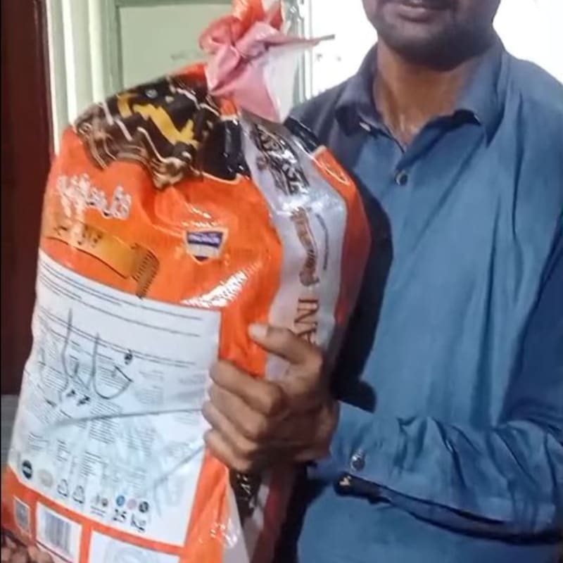 A man in a blue shirt holds a large bag of rice in the foreground, with a blurry indoor background.