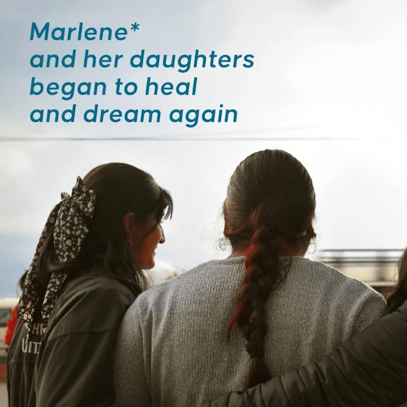 Three women, two of whom are looking at each other, are embraced in the foreground with a cloudy sky in the background.