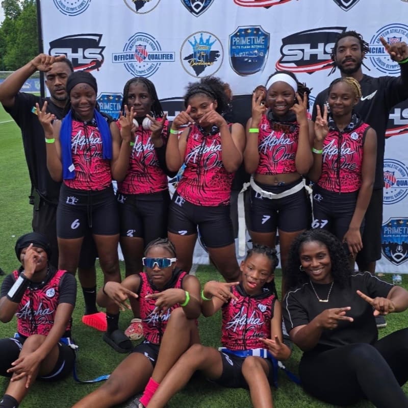 A group of young girls in matching pink and black athletic wear pose for a photo on a grassy field in front of a banner, with two adults standing behind them.