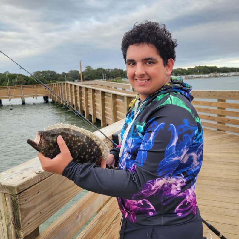 A young man in a colorful hoodie proudly displays a flatfish he caught on a wooden pier with a cloudy sky and water in the background.