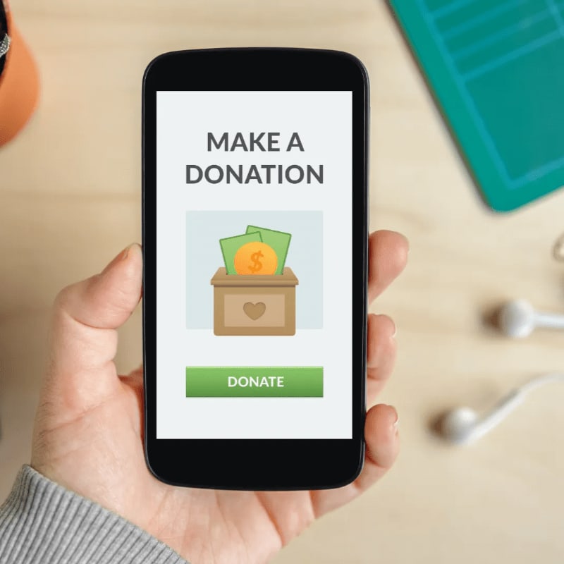 A hand holds a smartphone displaying a donation app with a wooden donation box and money, while a pair of white earbuds rests on a wooden desk in the background.