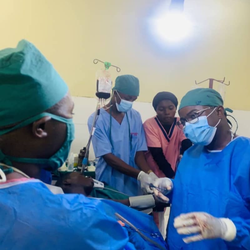 In the foreground, surgeons in blue scrubs and masks perform a procedure, while in the background, medical equipment and other staff are visible in a brightly lit room.