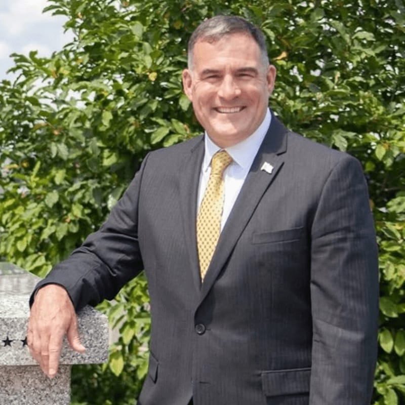 A smiling man in a suit leans on a stone structure in front of lush green foliage.