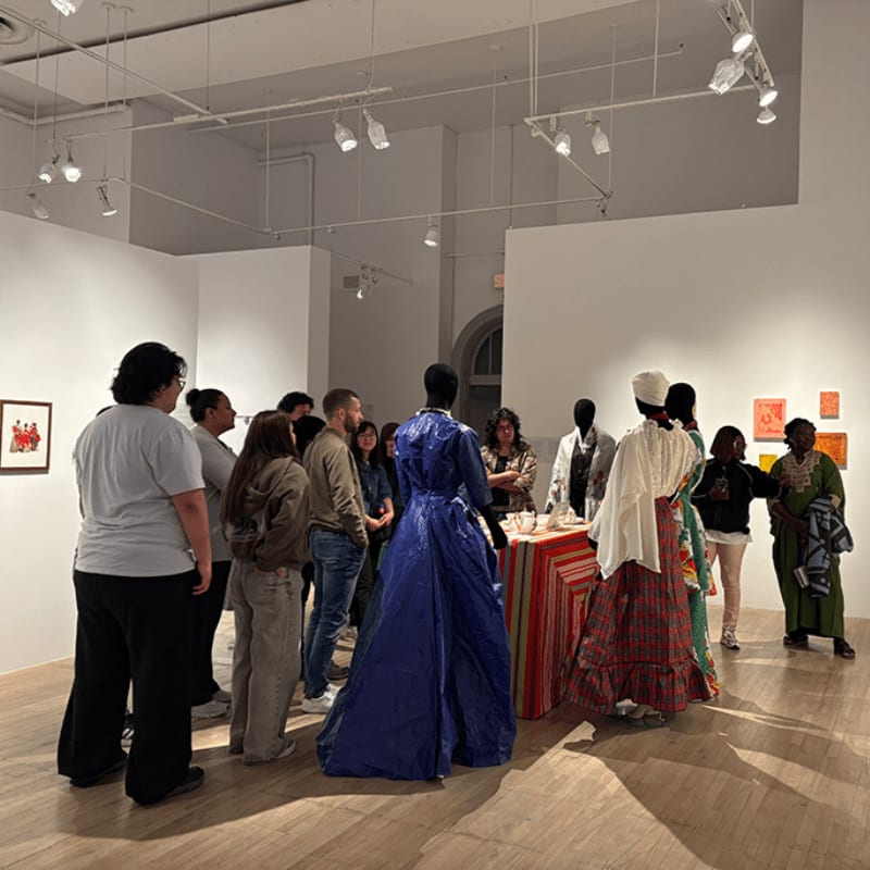 A group of people observe mannequins dressed in historical attire in a brightly lit art gallery with white walls and wooden floors.