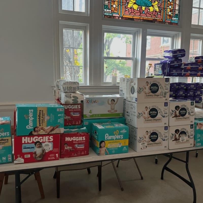 Boxes of diapers are stacked on tables in front of large windows with stained glass above.