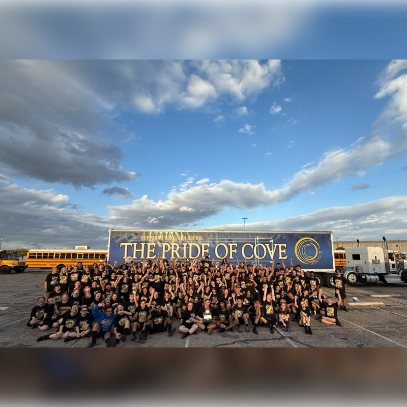 A large group of people in black t-shirts pose in front of a banner that reads "THE PRIDE OF COVE" with school buses and a semi-truck in the background under a cloudy sky.