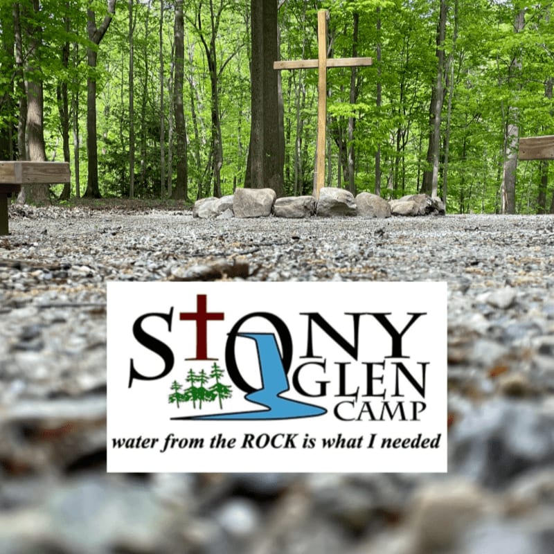 A wooden cross stands in a clearing surrounded by trees, with a logo for Stony Glen Camp in the foreground.