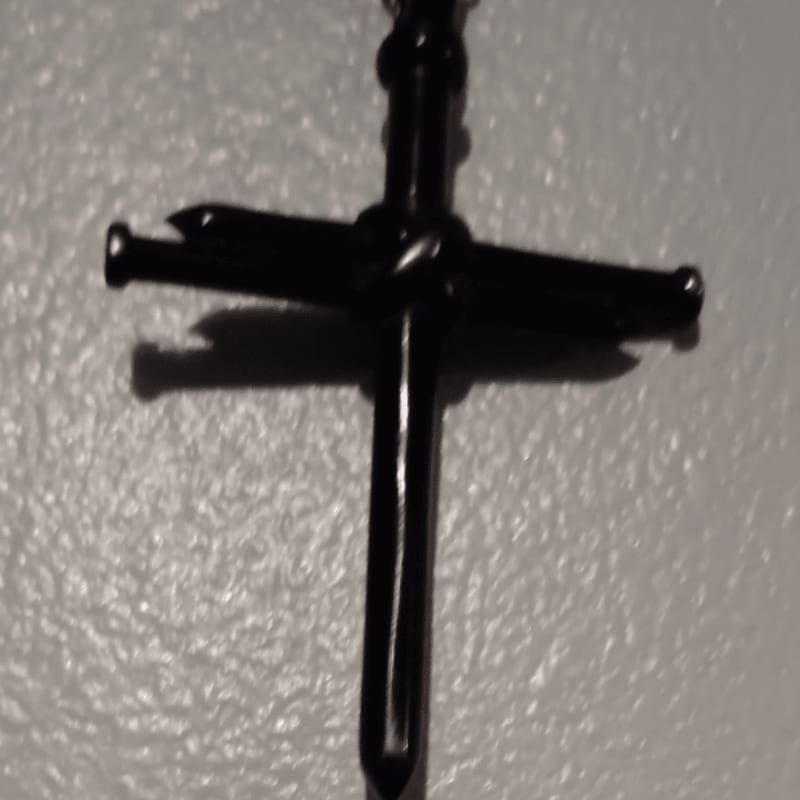 A dark, metallic cross hangs in front of a textured, light-colored wall.