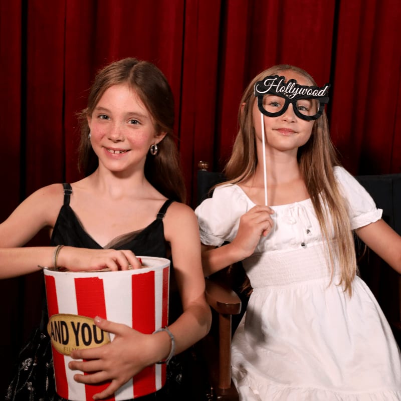 Two young girls pose with a popcorn bucket and a "Hollywood" prop in front of a red velvet curtain.