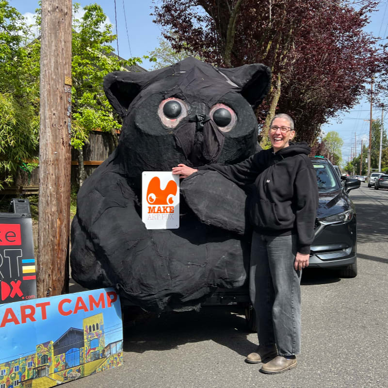 A person stands next to a large, black, owl-like sculpture holding a sign, with a street and parked cars visible in the background.
