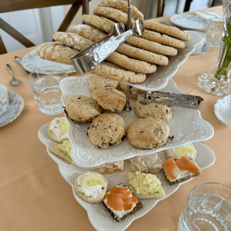 A tiered stand overflowing with cookies and small sandwiches sits on a peach tablecloth, with glasses of water and teacups in the background.