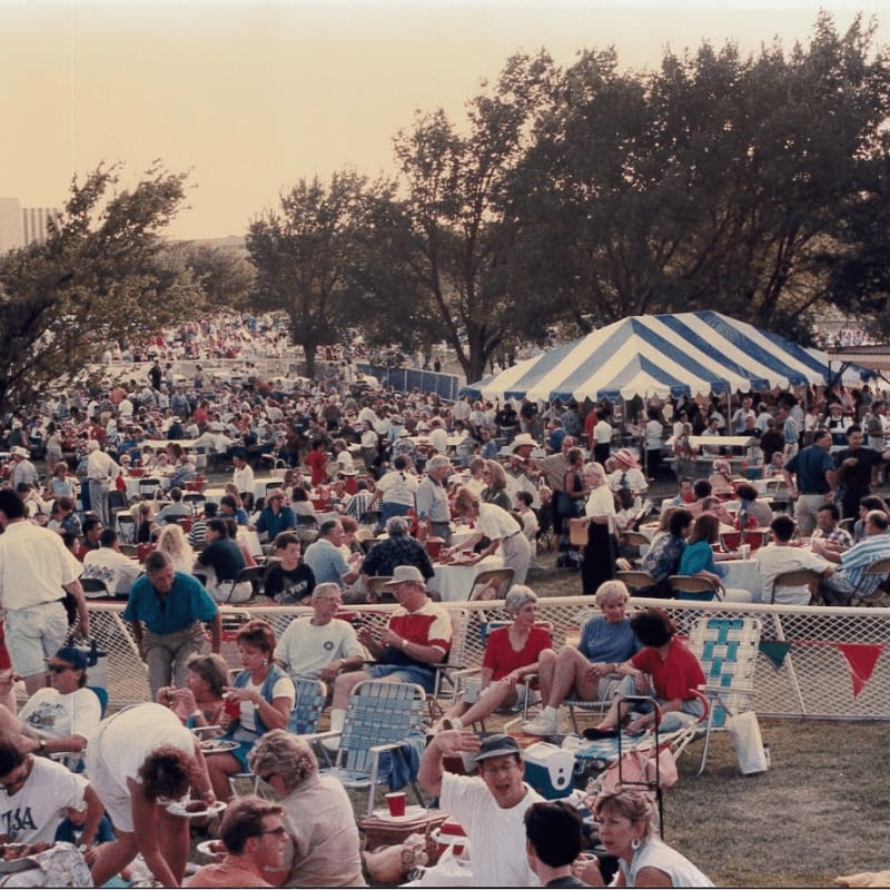 A large crowd enjoys a picnic on a grassy field in front of a striped tent and trees under a hazy sky.