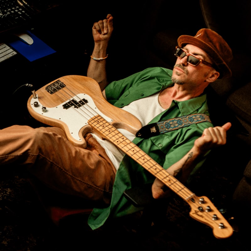 A man in a hat and sunglasses plays a bass guitar in the foreground, with a keyboard and computer mouse visible in the dimly lit background.