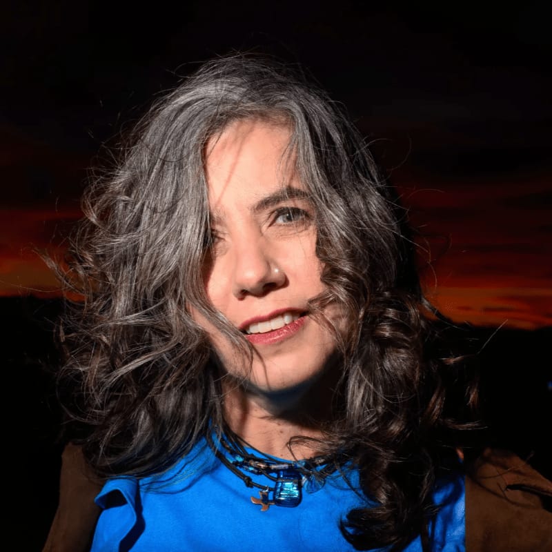 A woman with graying, curly hair and a blue shirt smiles at the camera with a warm, sunset-colored sky in the background.