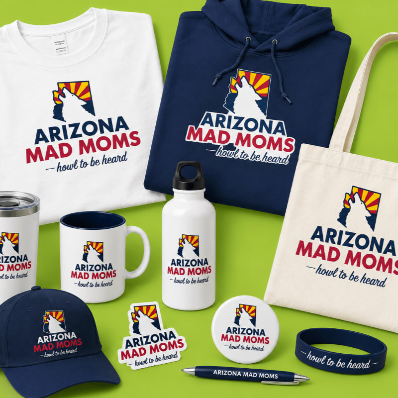 In the foreground, a collection of merchandise including a hat, mug, water bottle, stickers, a pin, a pen, and a wristband are displayed, while in the background, a white t-shirt and a navy hoodie, all featuring the "Arizona Mad Moms" logo, are laid out against a bright green surface.