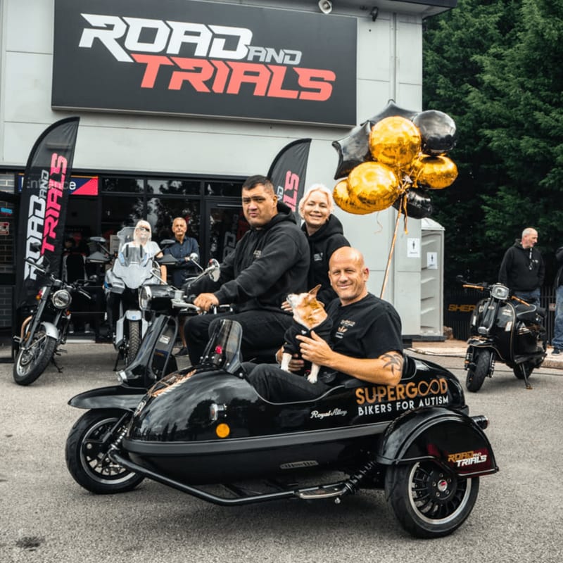 Three people and a dog sit in a black motorcycle sidecar in the foreground, with a building displaying "ROAD and TRIALS" signage and several motorcycles in the background.
