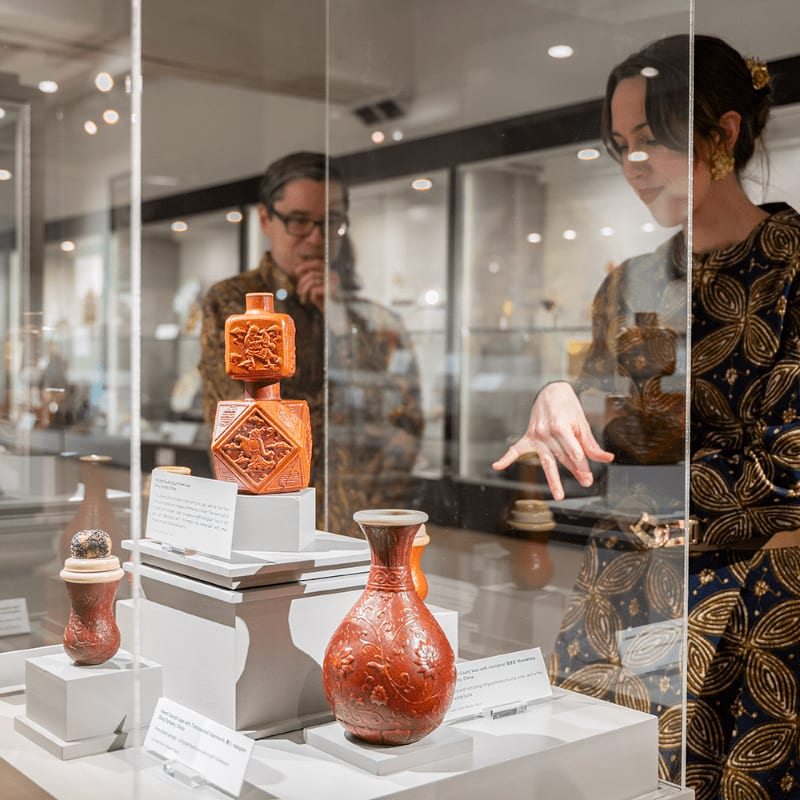 In the foreground, a woman in a patterned dress points to a red ceramic vase and carved wooden objects displayed in a glass case, while in the background, a man in a similar patterned shirt looks at other artifacts in a museum.