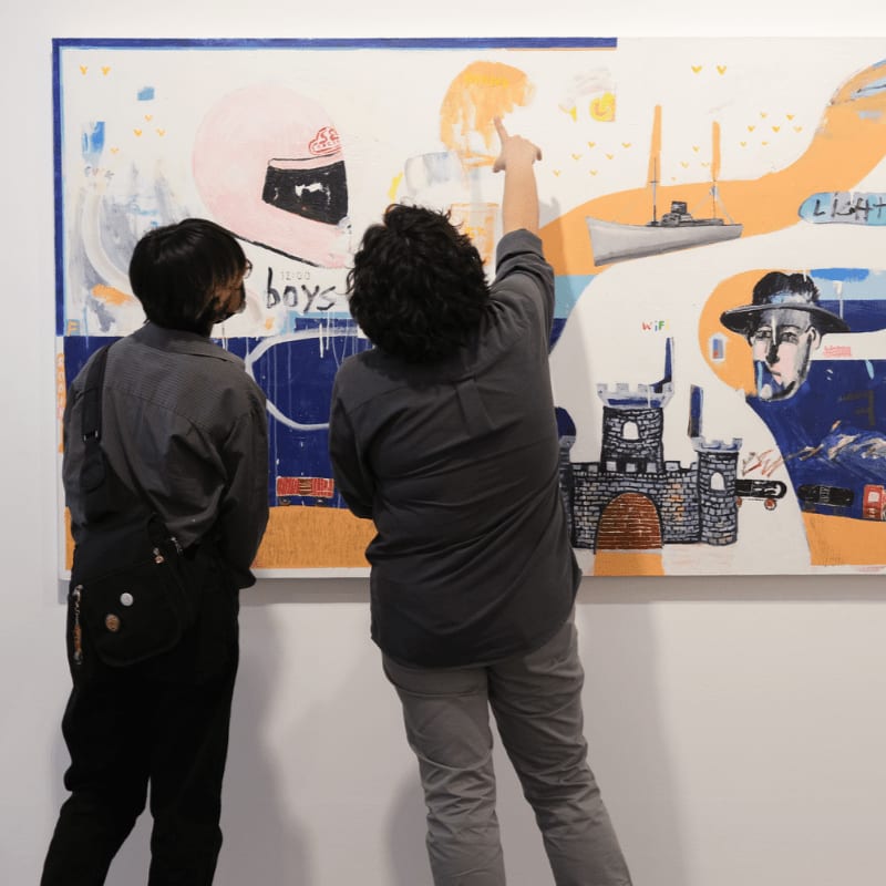 Two people stand before a colorful abstract painting featuring a pink helmet, a castle, and a man's face, with one person pointing at the artwork.