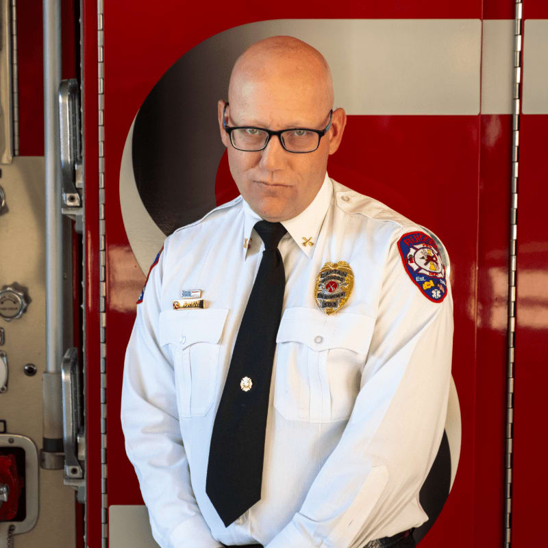 A bald man in a white uniform and black tie stands in front of a red fire truck.