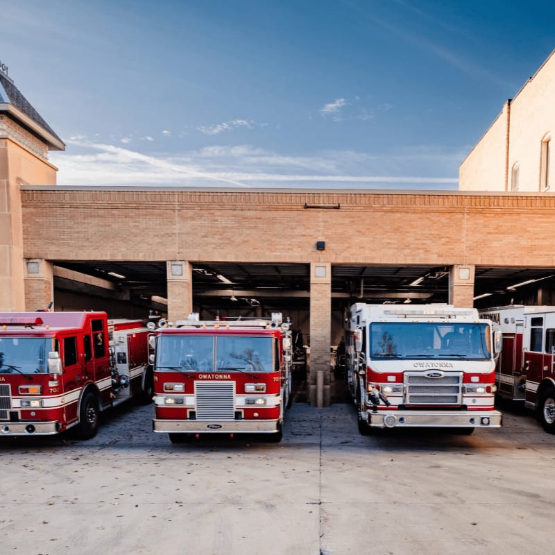 Four red fire trucks are parked in front of a brick fire station with a clear blue sky above.