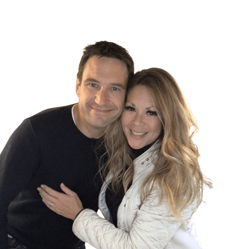 A smiling couple, a man in a dark sweater and a woman in a white jacket, pose closely together against a plain white background.