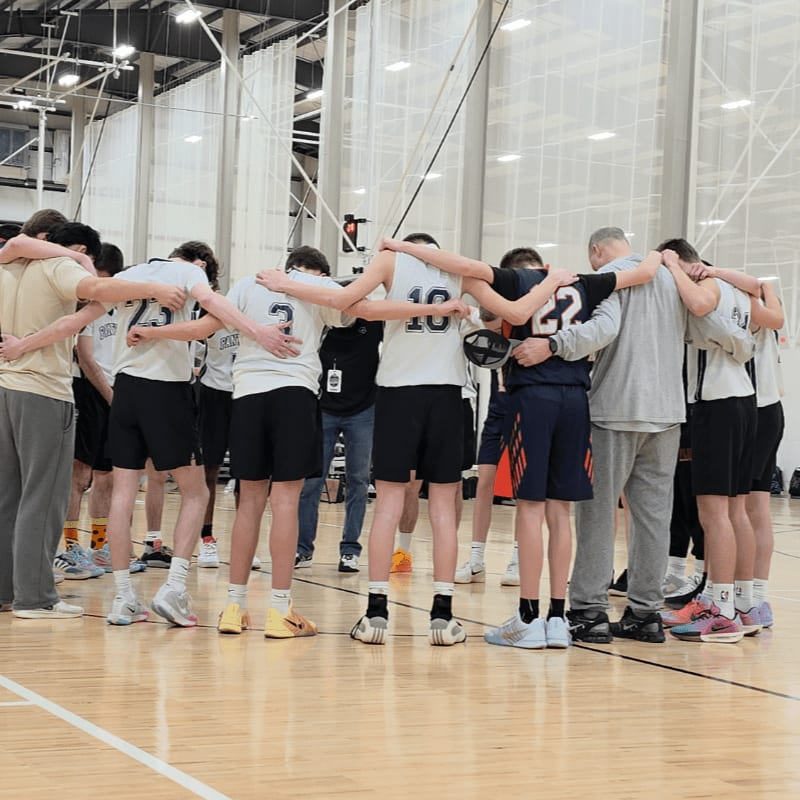 A group of young basketball players in uniform huddle together with their arms around each other on a polished wooden court, while spectators watch from the bleachers in the background.