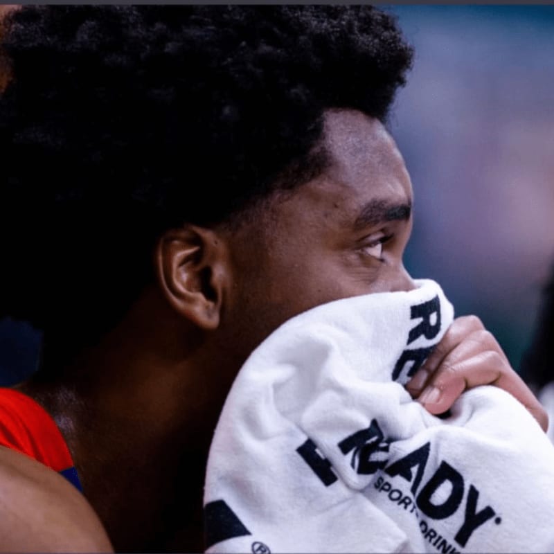 A basketball player in the foreground wipes his face with a white towel, while the blurred background suggests a game setting.