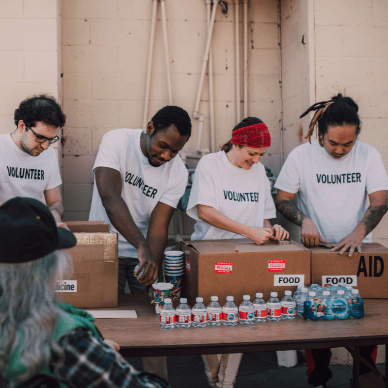 Volunteers in white t-shirts pack boxes of food and aid on a table in front of a textured wall.