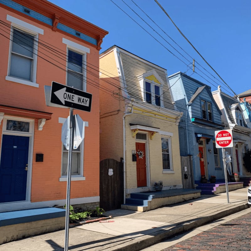 A bright orange house with a blue door stands in the foreground next to a yellow house, with a row of colorful houses and a clear blue sky in the background.
