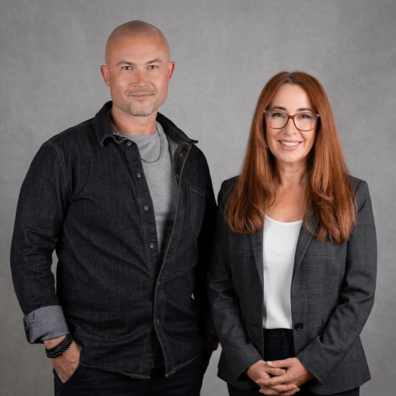 A bald man in a denim shirt and a woman with red hair in a blazer stand against a gray, textured background.