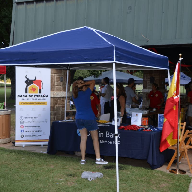 A blue tent with a "Casa de España" banner is set up on a grassy area, with people milling around in the background under a pavilion.