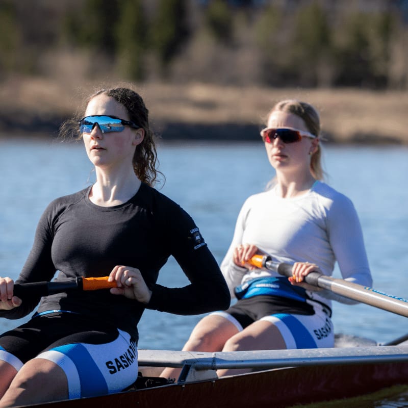 Two young women in athletic wear row a boat on a calm body of water, with trees and a clear sky visible in the background.