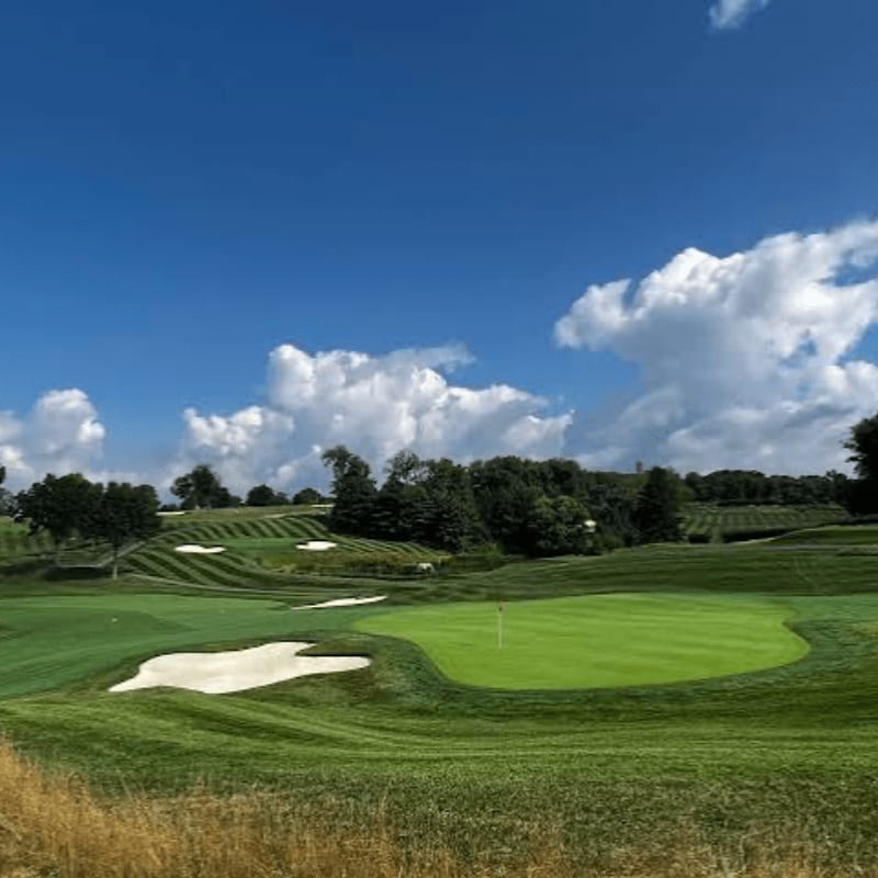 A manicured golf course green with sand traps in the foreground is set against a backdrop of rolling hills and a bright blue sky with fluffy white clouds.