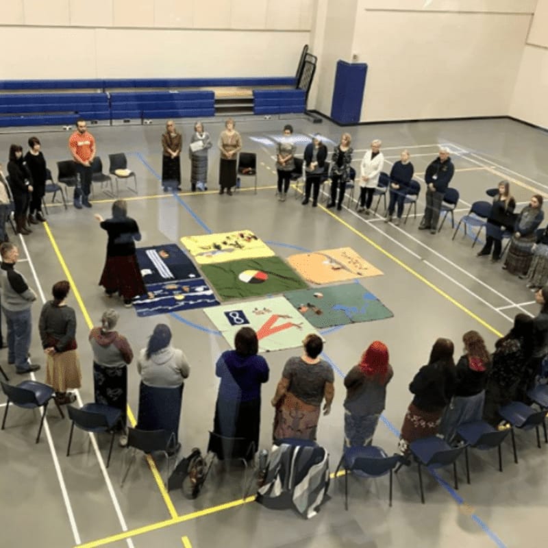 A group of people stand in a circle on a gymnasium floor around a display of colorful quilts, with bleachers visible in the background.