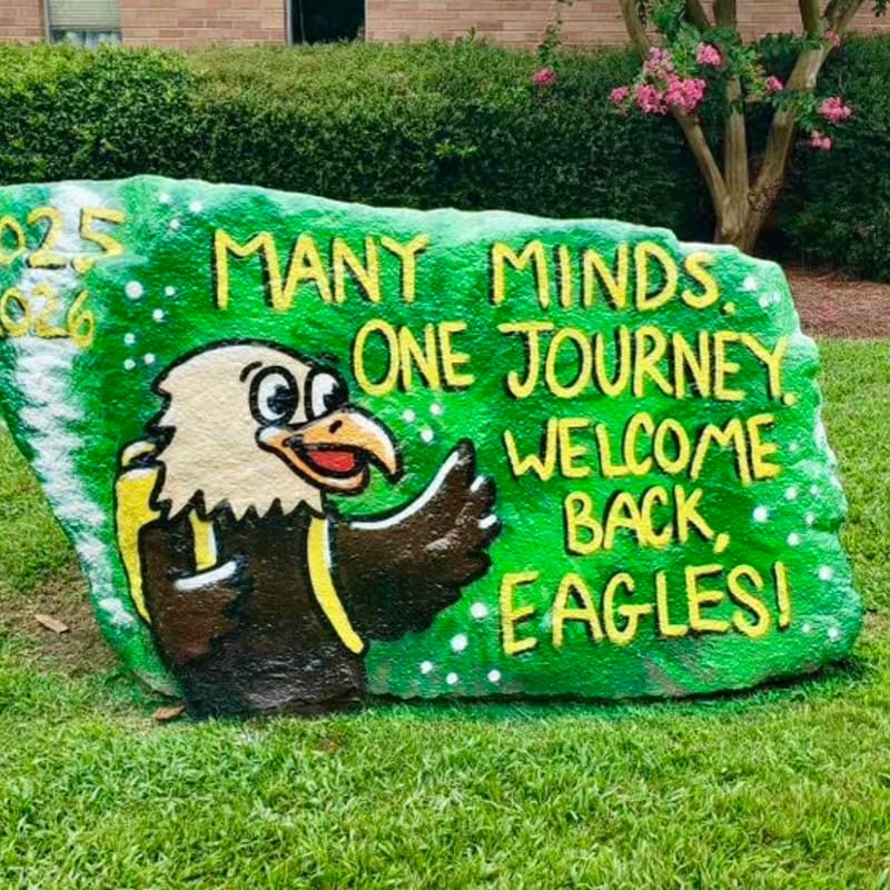 A painted rock with an eagle mascot and the words "Many Minds. One Journey. Welcome Back, Eagles!" sits on green grass in front of a building and a tree with pink flowers.