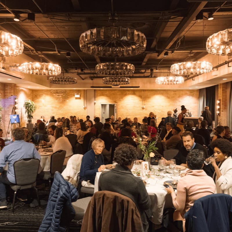 People are seated at tables in a dimly lit banquet hall with ornate chandeliers hanging from the ceiling.