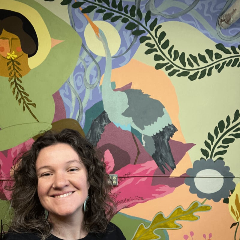 A smiling woman with curly brown hair is in the foreground, with a colorful mural of a heron and abstract nature elements in the background.