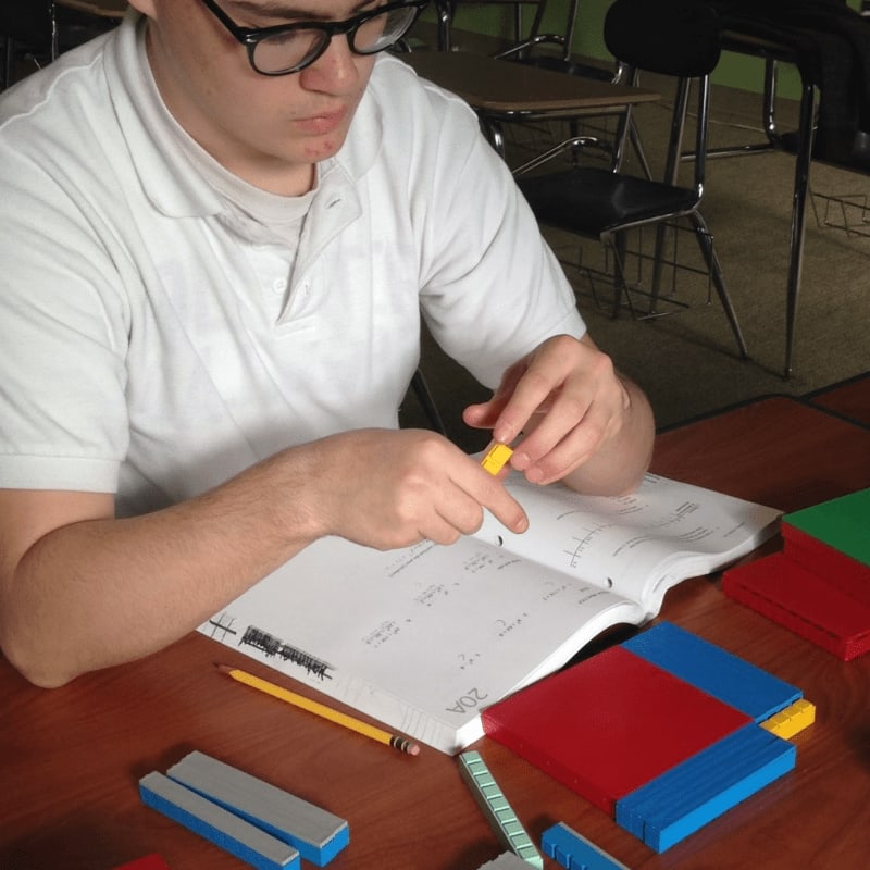 A student in a white shirt and glasses works with colorful blocks and a notebook at a desk in a classroom.