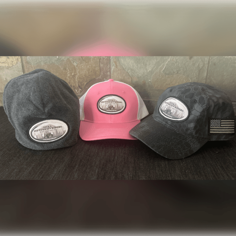 Three hats, one gray fleece, one pink and white trucker, and one black camouflage, sit on a dark surface in front of a stone wall.