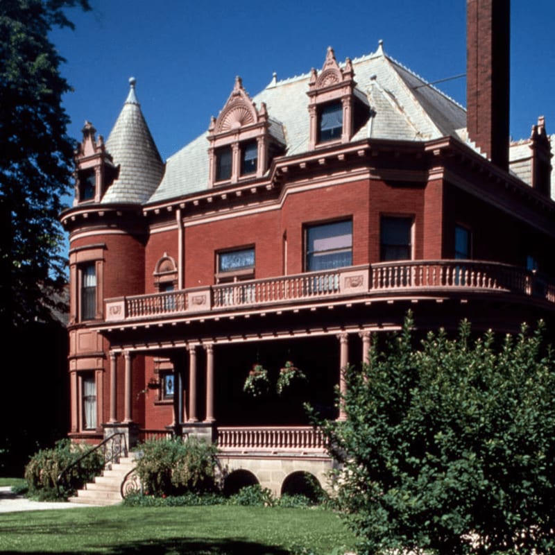A grand red brick Victorian mansion with a wraparound porch and turret stands against a clear blue sky, surrounded by lush green foliage.