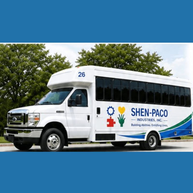 A white Shen-Paco Industries bus with colorful logos is parked in front of a line of green trees under a blue sky.