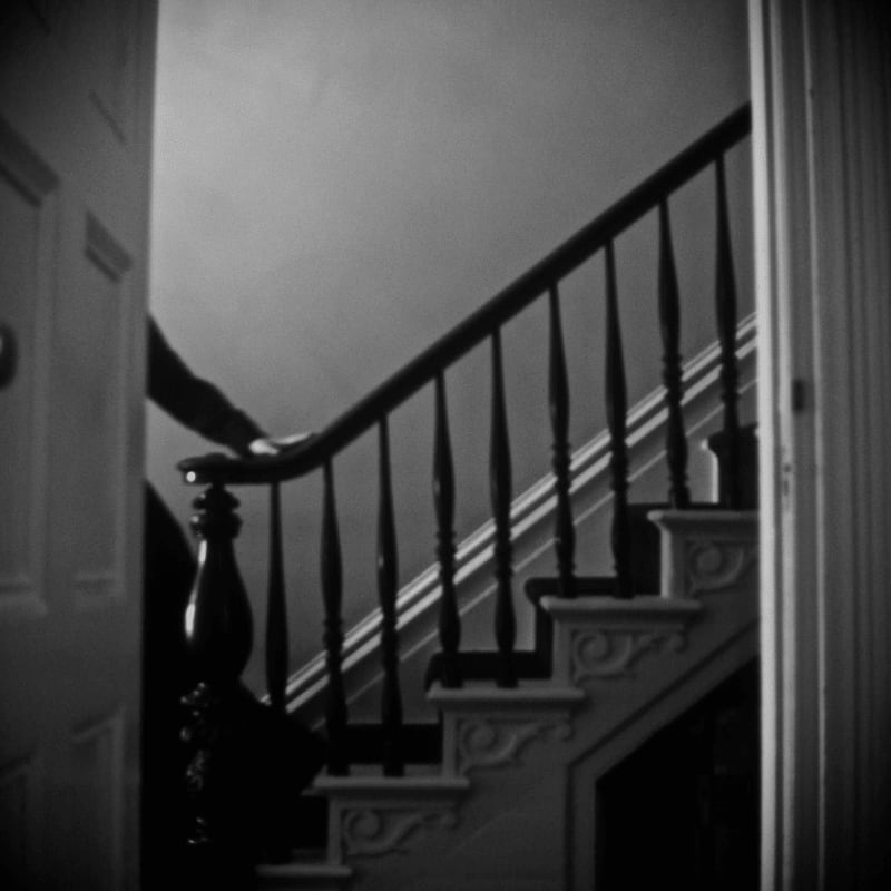 A hand rests on a dark wooden banister as it ascends a dimly lit staircase, framed by open doorways.