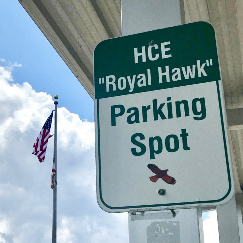 A green parking sign for "Royal Hawk" is in the foreground, with an American flag waving in the background under a cloudy sky.