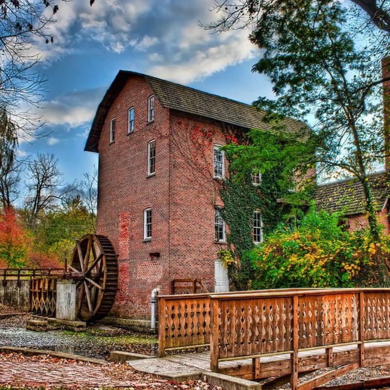 A rustic wooden bridge in the foreground leads to an old brick mill with a water wheel, surrounded by autumn foliage under a cloudy sky.