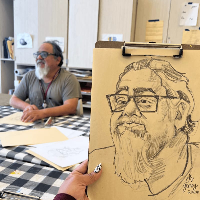 A sketch of a man with glasses and a beard is held in the foreground, while the man himself sits at a table in the background.